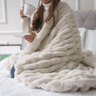 Person wrapped in a cozy beige blanket holding a cup on a bed.